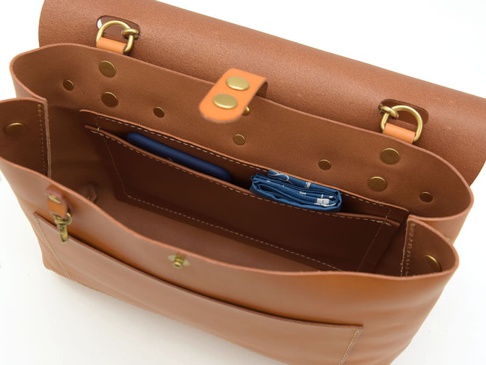 棕色皮革雙扣兩用側背包，內部結構可見手機隔層，另有藍色印花物品。包款使用黃銅色金屬釦具與鉚釘，精緻縫線。