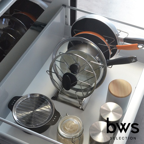 伸縮鍋具收納架，金屬材質，可收納鍋子與鍋蓋，整體呈現銀色調。架上放置著一個銀色鍋子，鍋蓋架上立著一個玻璃鍋蓋，鍋蓋上有兩個黑色把手。旁邊還有一個帶有橘色把手的黑色平底鍋。