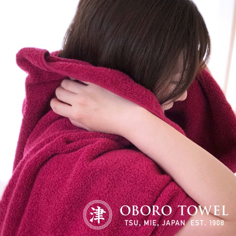 深紅色細纖浴巾，質地柔軟，一位女性正用浴巾擦拭頭髮，浴巾上有「津 OBOBO TOWEL TSU, MIE, JAPAN EST. 1908」字樣。