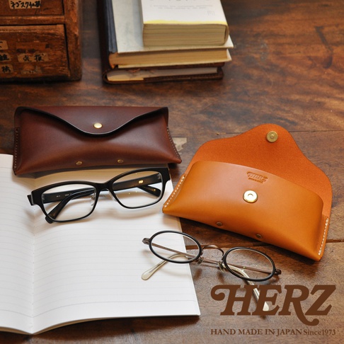 皮革眼鏡收納盒，有深棕色和淺棕色兩款，搭配黑框眼鏡和圓框眼鏡，以及一本筆記本，背景是木質桌面和書籍。