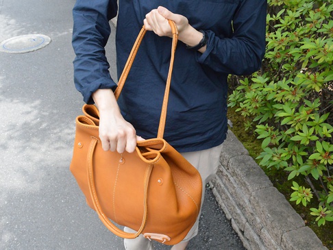 一位女士手持一個棕色皮革托特包，包身有縫線細節，搭配深藍色上衣和卡其色褲子，背景為綠色植物和石牆。