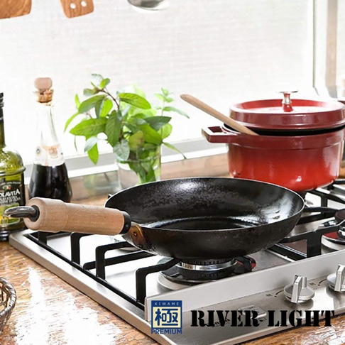 黑色不易生鏽平底鍋，鍋身呈現霧面質感，配有木製手柄，放置於不鏽鋼爐架上。背景有綠色植物和紅色鑄鐵鍋。