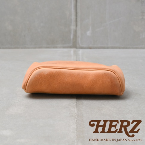 HERZ 品牌手工製作皮革包，側邊呈現圓潤飽滿的曲線，背景為灰色水泥牆面與地板，右下角有 HERZ 品牌 Logo 和「HAND MADE IN JAPAN Since1973」字樣。