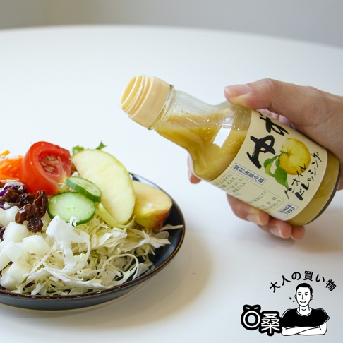 手持玻璃瓶裝無油柚子風味沙拉醬，醬汁呈淡黃色，瓶身有日文標籤，顯示柚子圖案。沙拉醬正淋在裝有生菜、番茄、小黃瓜、蘋果片和葡萄乾的深色沙拉碗中。