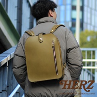 橄欖綠皮革製成，方形簡約設計的兩用後背包，搭配銀色拉鍊和金屬扣環，展示於穿著灰色外套的男性背後。