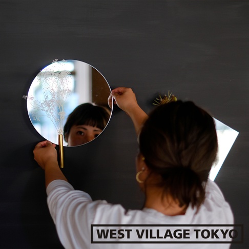 壁掛鏡面漂浮一輪花器，圓形鏡面，裝有細緻白色乾燥花束，搭配金色圓筒狀試管花瓶，懸掛於深色牆面上，鏡中映照出一位女性的臉龐。