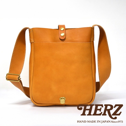日本HERZ職人手作皮革軟皮直式斜背包，淺棕色調，設有雙鈕釦翻蓋和外層口袋，配有可調節長度的肩帶，下方有一個黃銅色鎖扣。