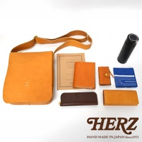 左側為駝色皮革斜背包，上方擺放著一本筆記本、兩個皮革皮夾，下方有咖啡色長夾及一個駝色皮夾，最右邊為黑色保溫瓶和藍色手帕，下方壓印有HERZ品牌標誌及HAND MADE IN JAPAN Since1973字樣。