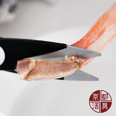不鏽鋼萬用料理剪刀正在剪切一塊蟹腳，剪刀刀刃為銀色金屬，手柄為黑色。