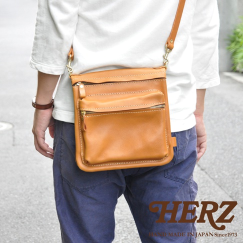 職人手作皮革翻轉雙色直式斜背包，採用天然皮革材質，呈現經典駝棕色，包身有兩個拉鍊前袋，搭配金屬拉鍊與縫線細節，斜背帶為可調整式，適合日常攜帶。