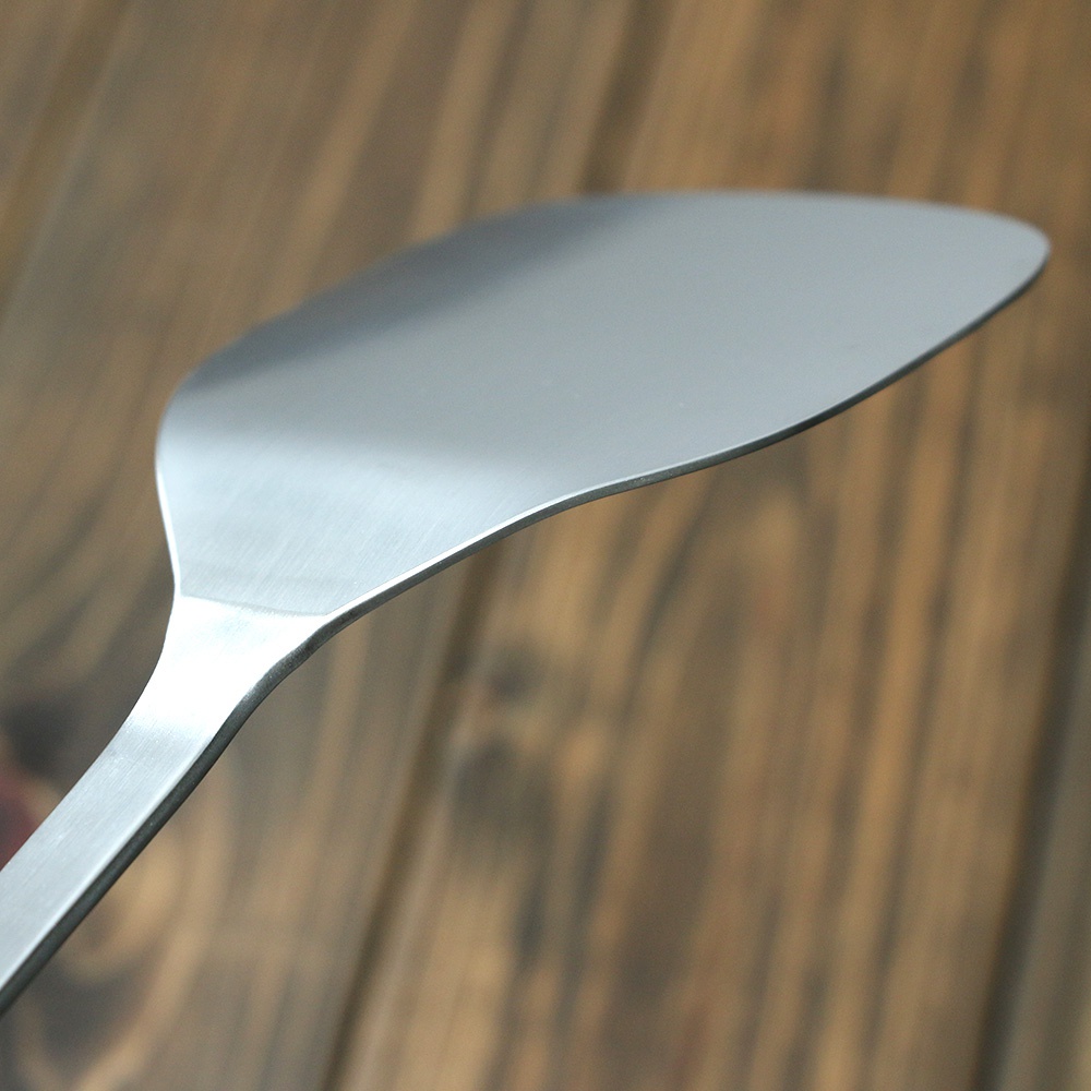 一個不鏽鋼萬用鍋鏟，其平坦的鏟頭呈扇形，邊緣略有弧度，整體呈現銀灰色金屬質感，與背景的木紋桌面形成對比。