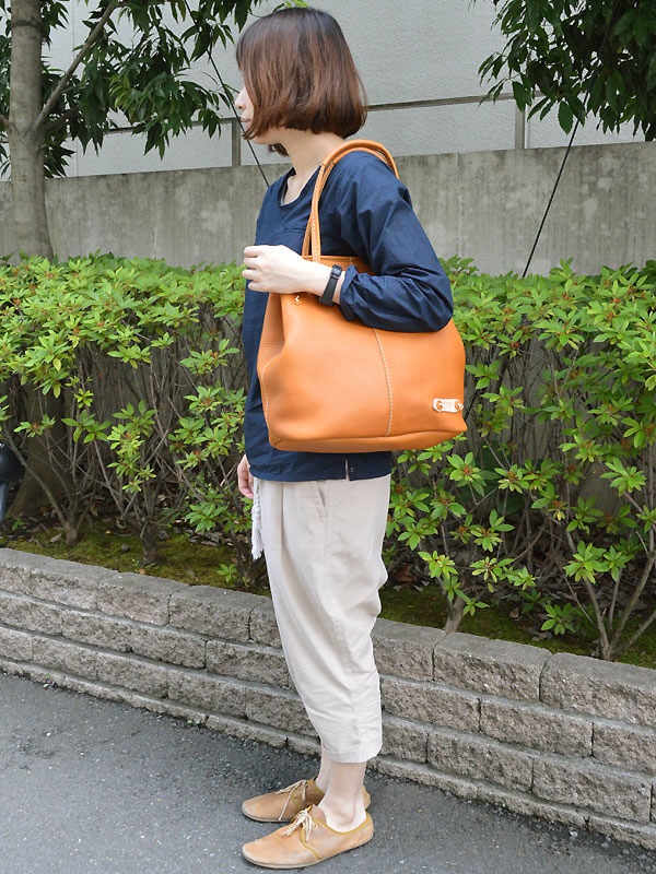 一位女性手提著一個棕褐色皮革托特包，包包有著明顯的縫線，底部有金屬扣飾。