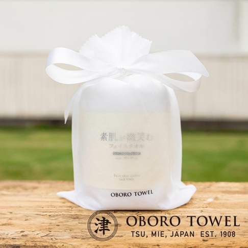 白色束口袋裝著一條印有日文「素肌、微笑」與英文「bare skin smiles FACE TOWEL」的毛巾，束口袋頂部綁著白色緞帶，放置於木質桌面，背景為模糊的綠色草地與建築。