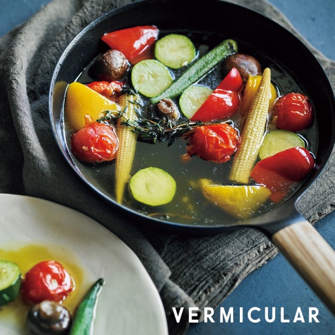 日本製黑色琺瑯鑄鐵平底深鍋，內含煮熟的番茄、黃甜椒、櫛瓜、玉米筍、秋葵及香草。