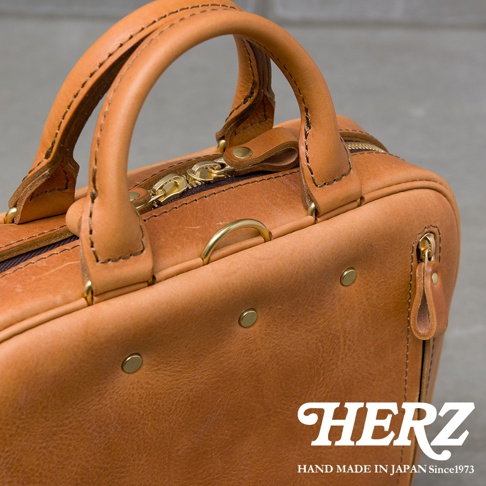 職人手作皮革公事包，近距離特寫，包含提把、拉鍊和金屬細節，背景為灰色水泥牆，右下角有品牌標誌 HERZ HAND MADE IN JAPAN Since1973