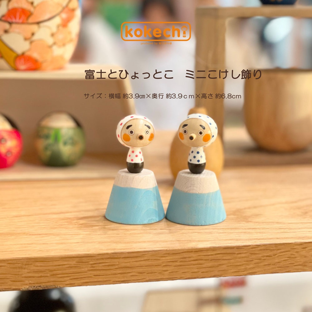 富士與火男木芥子開運擺飾組合。兩個木製達摩娃娃，分別穿著白色帶紅點和藍點的衣服，臉部有圓形紅臉頰和開口嘴巴。娃娃們站立在淺藍色和原木色的富士山形狀底座上。