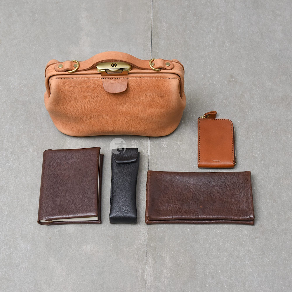 日本HERZ職人手作皮革Organ鎖孔式大開口肩背包M-7，搭配深棕色長夾、黑色筆套、棕色短夾及深棕色筆記本，置於灰色紋理地板上。