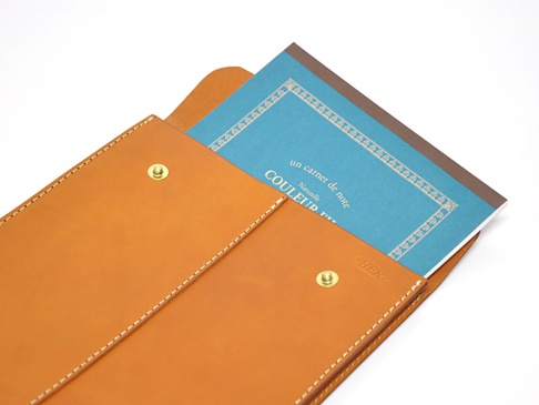 職人手作皮革信封式雙釦手拿袋，採用自然色皮革，搭配金色釦環，內含一本藍色封面筆記本，展現簡約質感。