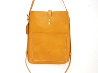 職人手作皮革散步直式單肩包，天然皮革材質，呈現溫潤的焦糖棕色。包身線條簡潔，正面設有一個方形貼袋，袋口以白色縫線勾勒，質感樸實。包蓋處有兩顆金屬卯釘，並以皮革搭扣固定。細長的皮革背帶垂掛，整體風格自然休閒。