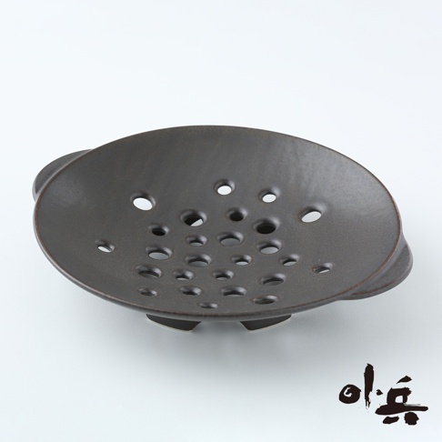 深棕色陶製多用蒸盤，盤面佈滿大小不一的透氣孔，盤緣有兩個小提把，盤底有三個支撐腳。