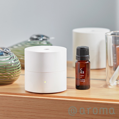 簡潔的白色圓柱形隨身精油香氛機，頂部中央有圓孔，側面下方有黃色指示燈。旁邊擺放著一瓶琥珀色玻璃瓶裝的精油，瓶身標有「C 03 Clean air CLEAN FOREST」字樣，瓶蓋為黑色。背景還有綠色條紋的玻璃裝飾品和白色圓筒。