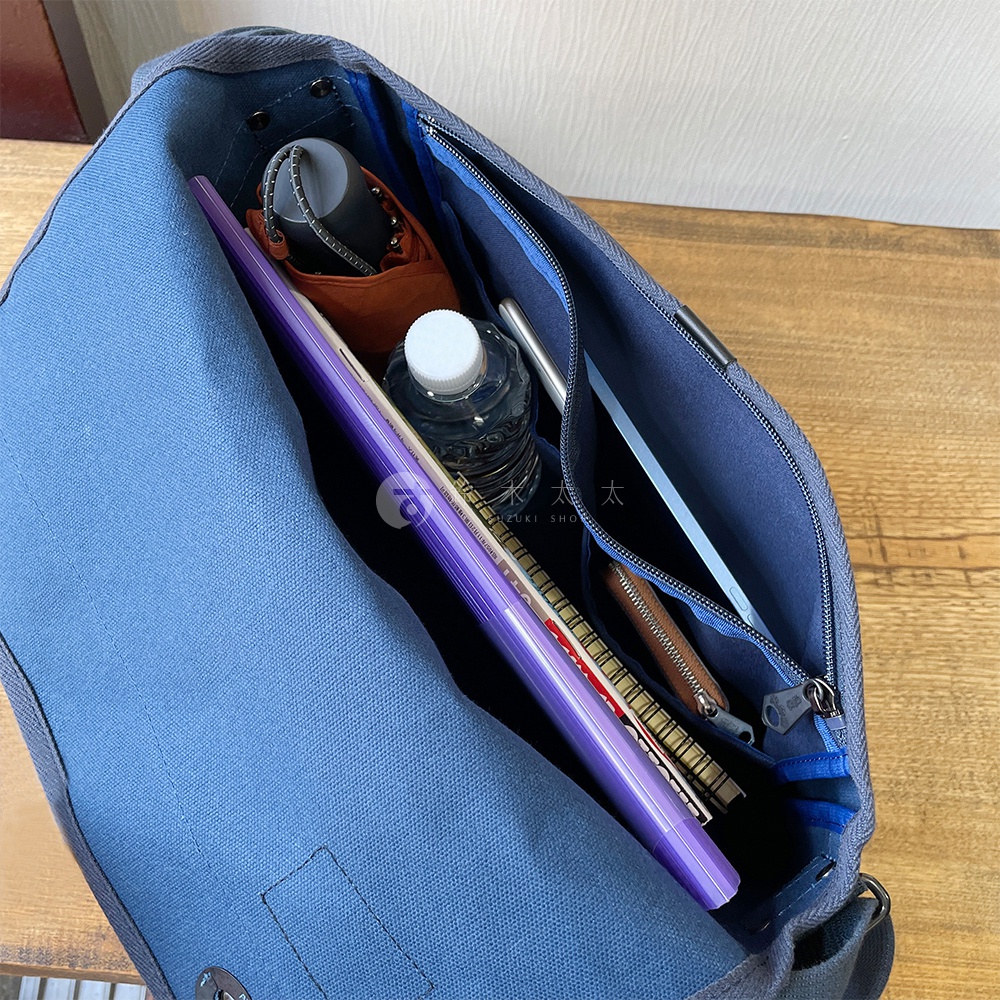 黑色帆布郵差包，內部展示，裝有紫色筆記本、水瓶、筆及其他小物，包包為藍色調。