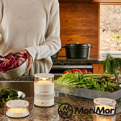 松本素白LASMO Speaker 燭光氣氛音響，白色圓柱形主體，頂部為透明圓形燈罩，內部有LED燈發出溫暖黃光。底部為白色喇叭網罩，印有MoriMori Logo。