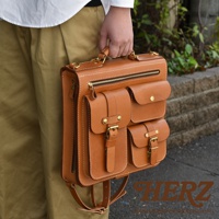 職人手作皮革三口袋三用後背包，採用天然皮革材質，呈現溫潤的淺棕色。包款設計有頂部提把、側邊拉鍊，正面則有兩個翻蓋式口袋及下方一個較大的翻蓋式口袋，皆配有金屬釦環及皮帶束帶，提供多樣化的收納與攜帶方式。