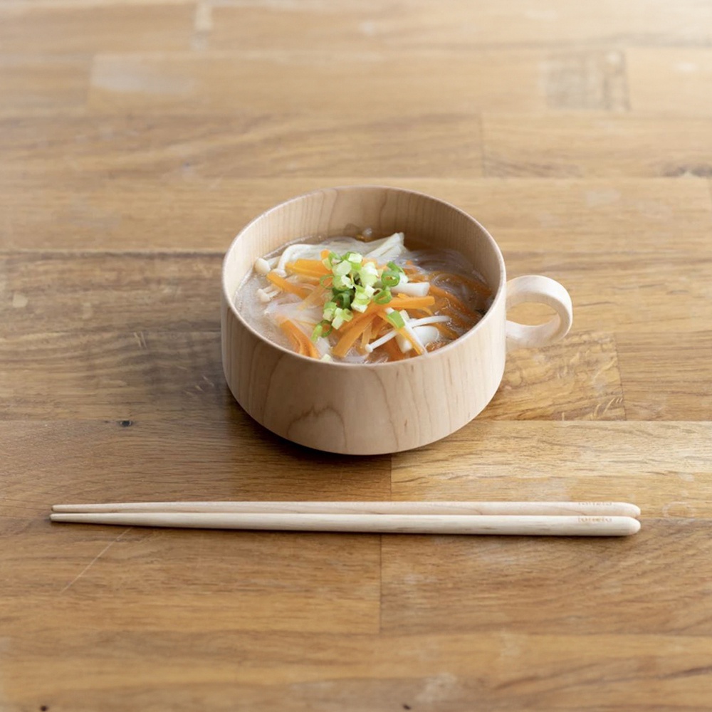 一雙淺色楓木圓筷，平放在木質桌面上，筷子旁是一個裝有湯麵、紅蘿蔔絲、金針菇和蔥花的木碗。