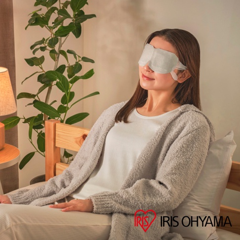 一位女性坐在室內舒適地放鬆，頭戴淺灰色 Iris Ohyama 品牌蒸氣眼罩，身穿柔軟的灰色針織外套，背景有溫暖的檯燈與盆栽，營造出居家放鬆的氛圍。