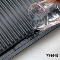 深灰色波浪紋烤盤，烤盤表面有條紋，一個透明玻璃量杯傾倒清水，水流進入烤盤的凹槽中。