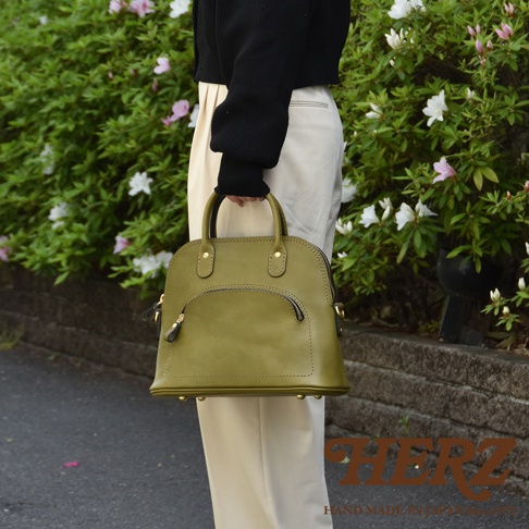 橄欖綠皮革貝殼造型手提包，配有頂部提把、拉鍊開合主袋及前方拉鍊口袋，底部有金屬腳釘。