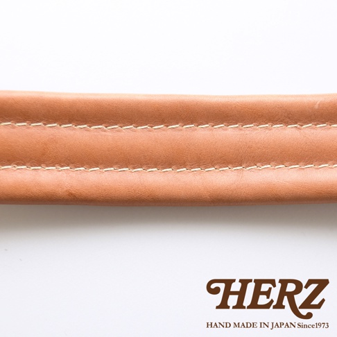 職人手作皮革加厚減壓肩背帶，呈現自然的淺棕色皮革紋理，搭配米白色縫線，質感細緻，並印有品牌HERZ及HAND MADE IN JAPAN Since1973字樣。