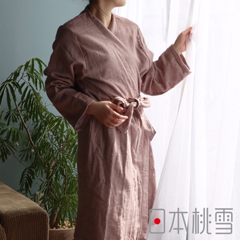 一位女性穿著一件粉色紗質浴袍，腰間繫著同色系的腰帶，手輕觸窗簾。