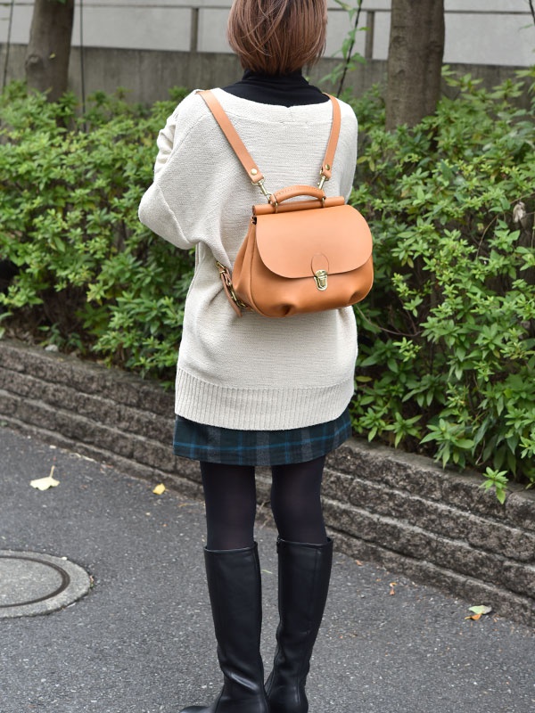 淺棕色皮革迷你後背包，帶有翻蓋和金色鎖扣，搭配米白色毛衣與格紋短裙。