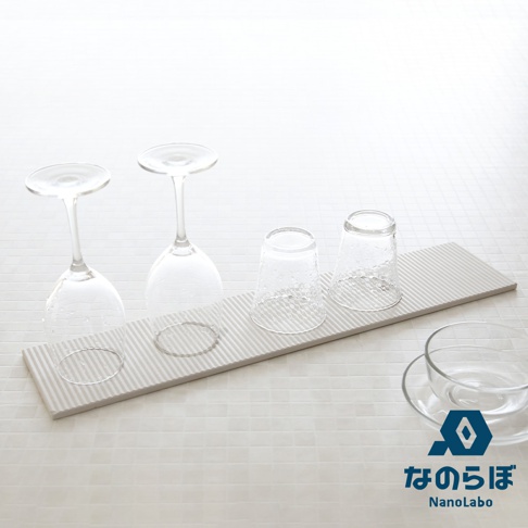 淺灰色長形珪藻土置物墊，表面有細長條紋，上面放置兩個倒置的透明玻璃酒杯和兩個直立的透明錘紋玻璃杯，右下角有透明玻璃碗和品牌 Logo。