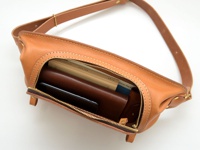 棕色皮革拉鍊旅行斜背包，主隔層打開，內有手機、皮夾和書籍。包包採用皮革材質，配有可調節的肩帶和黃銅色拉鍊。