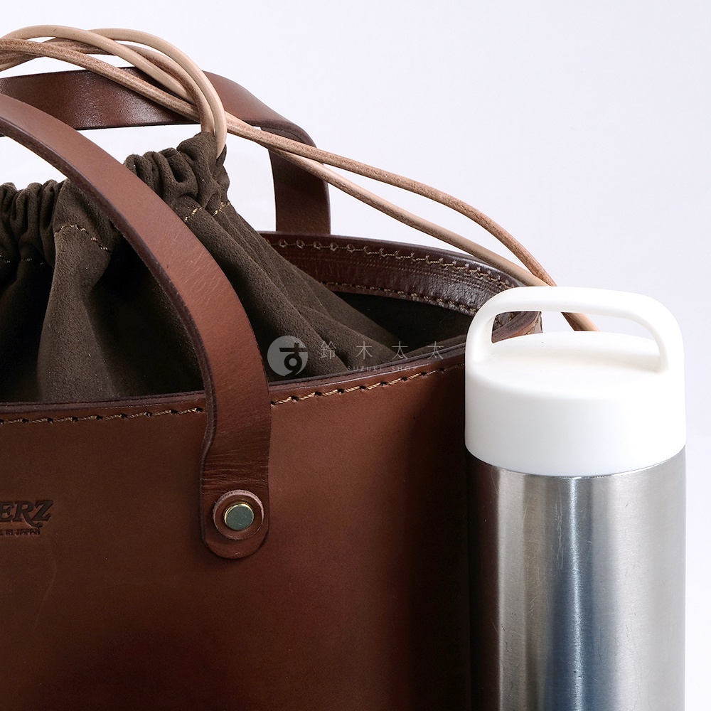 職人手作深棕色皮革束口手提包，內襯為深綠色麂皮，搭配兩條淺棕色皮革提把和束繩。包身一側有銀色金屬扣飾，右側放置一個白色瓶蓋的銀色保溫瓶。