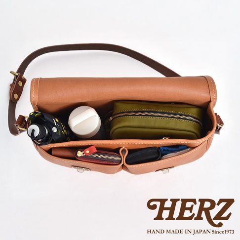 職人手作皮革傳統釣魚包，焦糖色皮革，包內裝有黑色雨傘、白色圓形小物、軍綠色化妝包、紅色拉鍊零錢包及黑色皮夾。