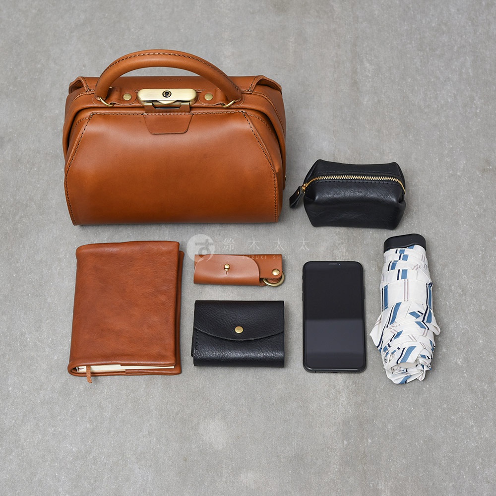 職人手作皮革Organ兩用口金手提包，呈現棕色皮革，搭配金色金屬扣環。一旁擺放著黑色皮革零錢包、棕色皮革鑰匙圈、黑色皮夾、手機，以及一把印有藍白幾何圖案的摺疊傘。