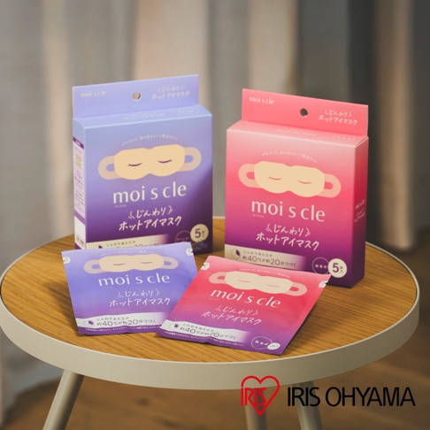 桌面上放置著兩盒與兩包 IRIS OHYAMA 的 moiscle 蒸氣眼罩，分別為紫色與粉紅色包裝。盒裝上清楚標示 5 枚入，單包裝則標註了約 40 度的溫熱感可持續約 20 分鐘的特色，背景為簡約溫暖的室內風格。