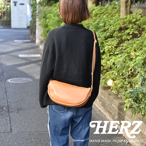 日本HERZ職人手作皮革半圓馬鞍包，由後方視角展示，模特兒身穿黑色上衣和牛仔褲，側背著一個棕色皮革製成的半圓形包款，包款上有HERZ品牌字樣和HAND MADE IN JAPAN Since1973的字樣。