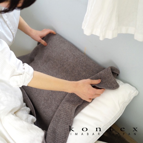 一條深棕色針織風純棉毛巾，質地柔軟，蓋在白色枕頭上。