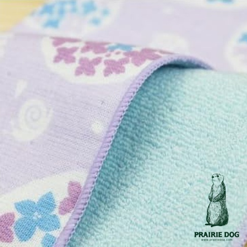一塊淡紫色印有可愛卡通蝸牛和花朵圖案的方巾，邊緣縫有淺紫色滾邊，下方露出淺藍色的毛巾布料，右下角有一隻戴著太陽眼鏡的土撥鼠插畫及PRAIRIE DOG字樣。