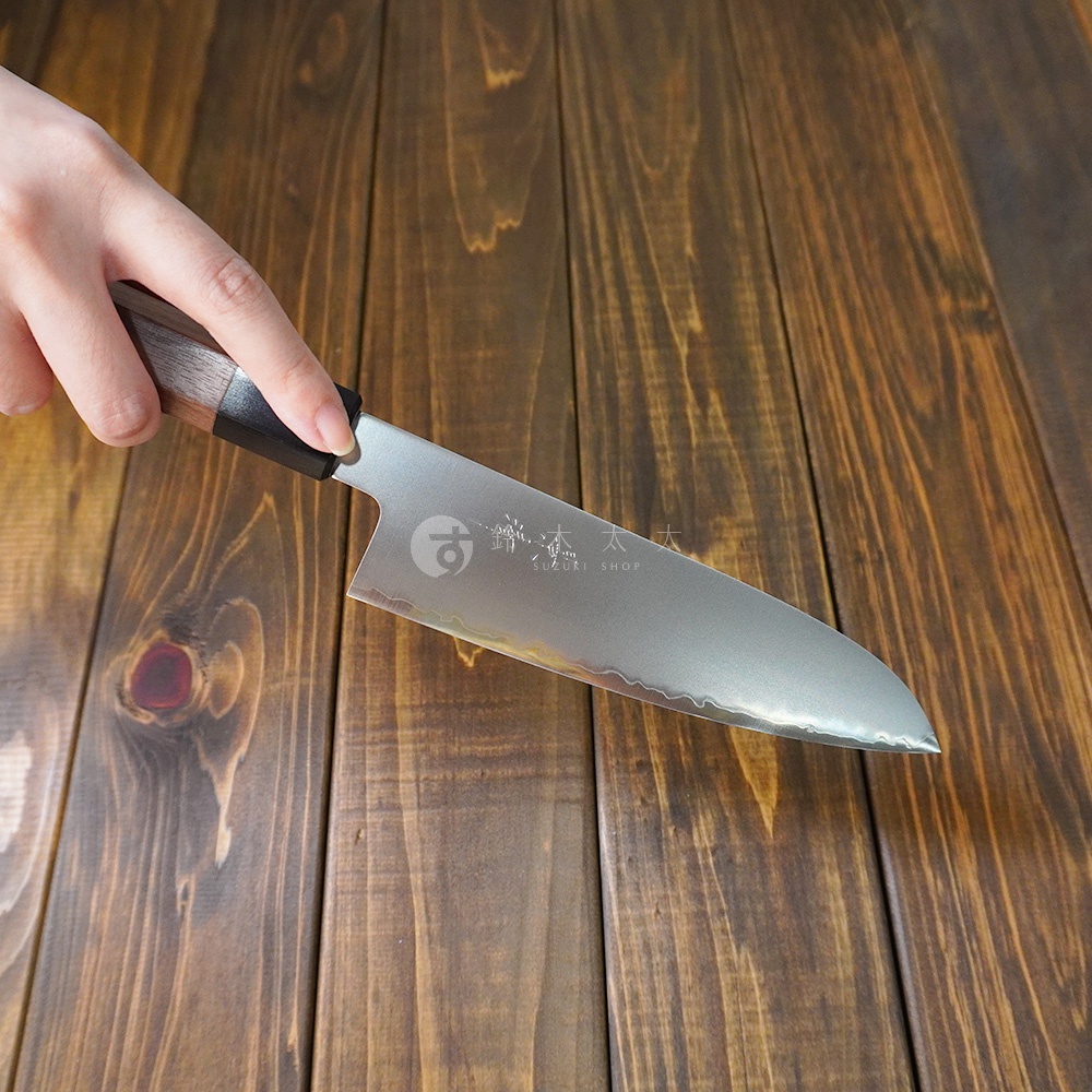 一只手握著一把銀色Chromax鋼三德刀，刀身上有波浪紋和品牌標誌。刀柄為深棕色胡桃木材質，與木紋桌面背景形成對比。