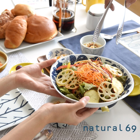 一雙手捧著一個白色陶瓷沙拉碗，碗內裝有豐富的沙拉，包含生菜、紅蘿蔔絲、甜椒、地瓜片和蓮藕片，碗邊有藍色花紋。背景有多種麵包、小碟和餐具。