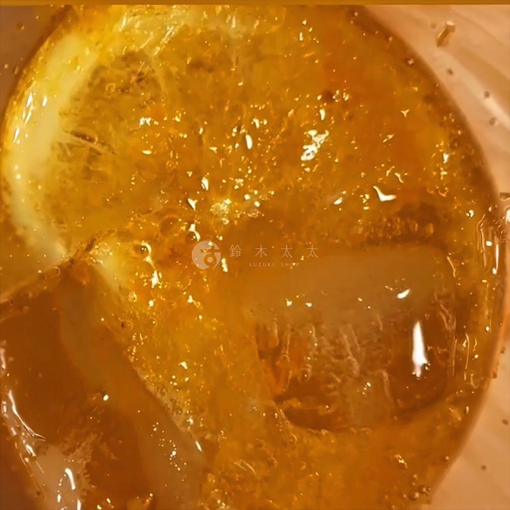 俯瞰視角呈現一杯冰鎮的廣島無添加手工熟成蜜柑檸檬釀，金黃色的液體中漂浮著一片新鮮檸檬與通透的冰塊，氣泡在杯中細膩流動，呈現出清爽沁涼的視覺質感，杯身中央印有鈴木太太的品牌標誌。