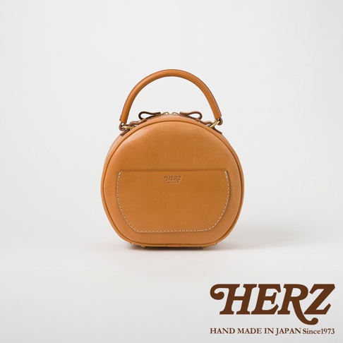 日本HERZ品牌手工製作的駝色皮革圓形兩用包，帶有前置口袋和HERZ LOGO。