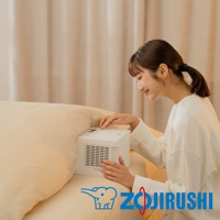 一位身穿白色毛衣的女性將手伸向一個白色多功能智慧烘乾機，該烘乾機放置在米色床墊上，背景是米色窗簾。