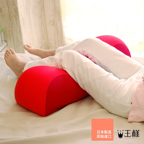 一對紅色半圓形膝下枕，置於床上，一人仰臥，雙腿彎曲，膝蓋分別放置在枕頭上。